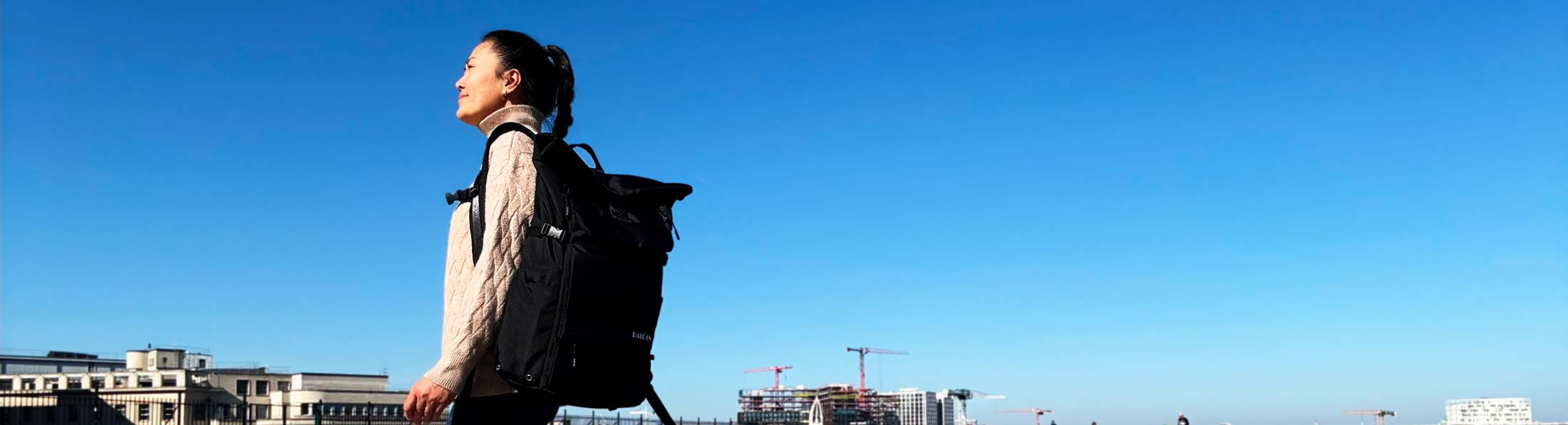 Junge Frau mit schwarzem Rolltop-Rucksack von BALÔ-ON geht durch einen großen Platz; im Hintergrund sind moderne Hochhäuser und Krane zu sehen.