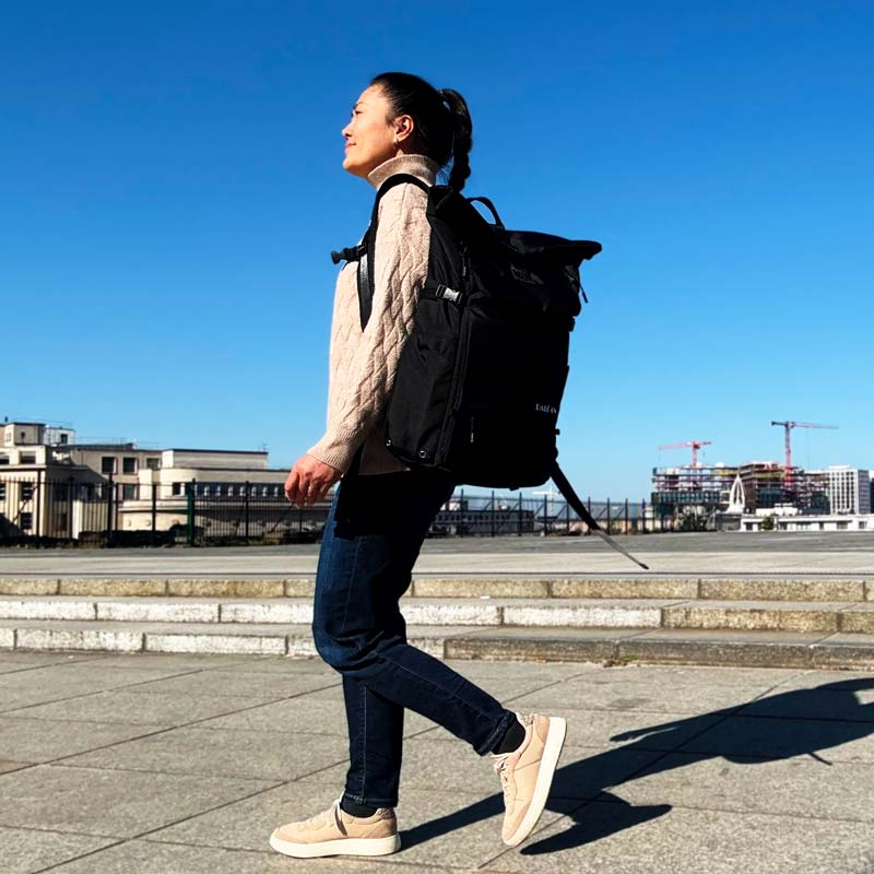 Junge Frau mit schwarzem Rolltop-Rucksack von BALÔ-ON geht durch einen großen Platz; im Hintergrund sind moderne Hochhäuser zu sehen.