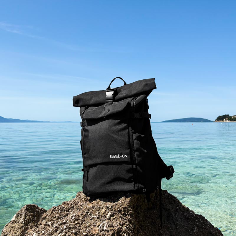 Rucksack steht auf einem Felsen am Meer, im Hintergrund ist das Meer und blauer Himmel zu sehen.