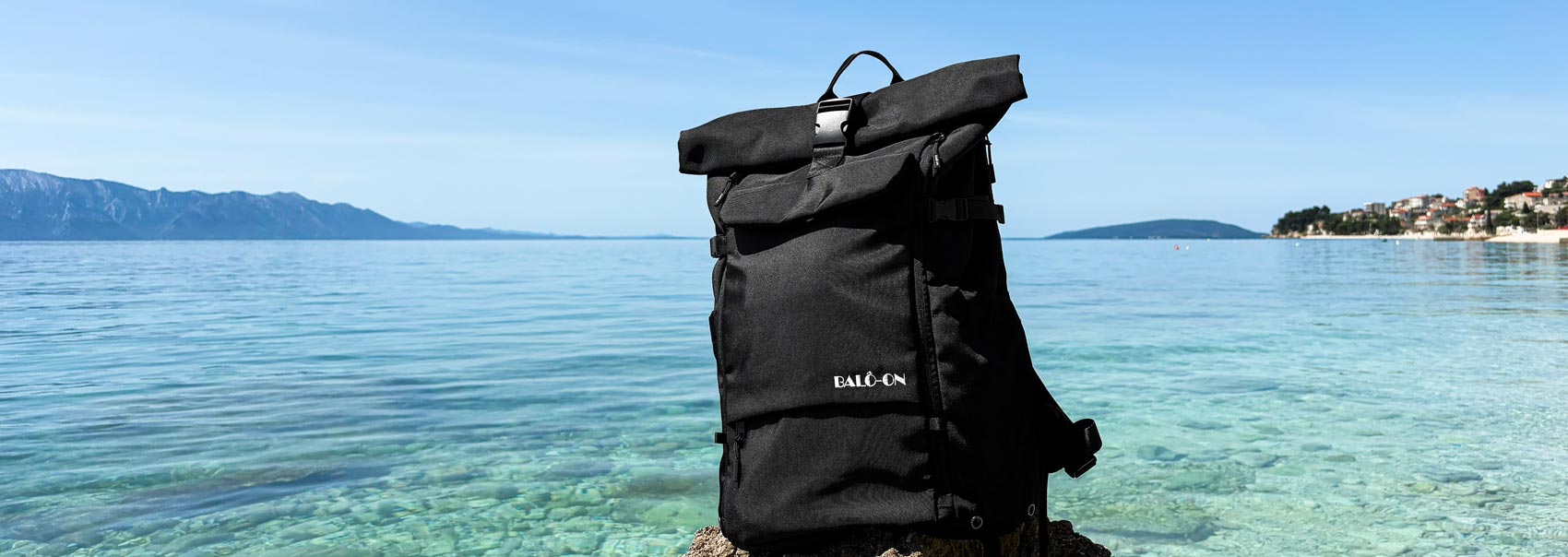Schwarzer BALÔ-ON Rucksack steht auf einem Felsen am Meer, im Hintergrund ist das Meer und blauer Himmel zu sehen. Bild im Querformat.