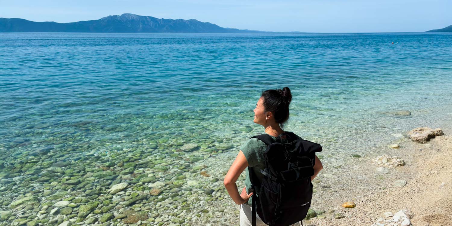 Junge Frau mit schwarzem Rucksack der Marke BALÔ-ON steht am Strand, blickt in die Ferne; türkisfarbenes Wasser und Berge im Hintergrund.