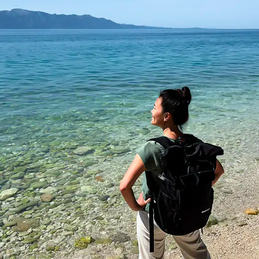 Junge Frau mit schwarzem Rucksack der Marke BALÔ-ON steht am Strand, blickt in die Ferne; türkisfarbenes Wasser und Berge im Hintergrund.