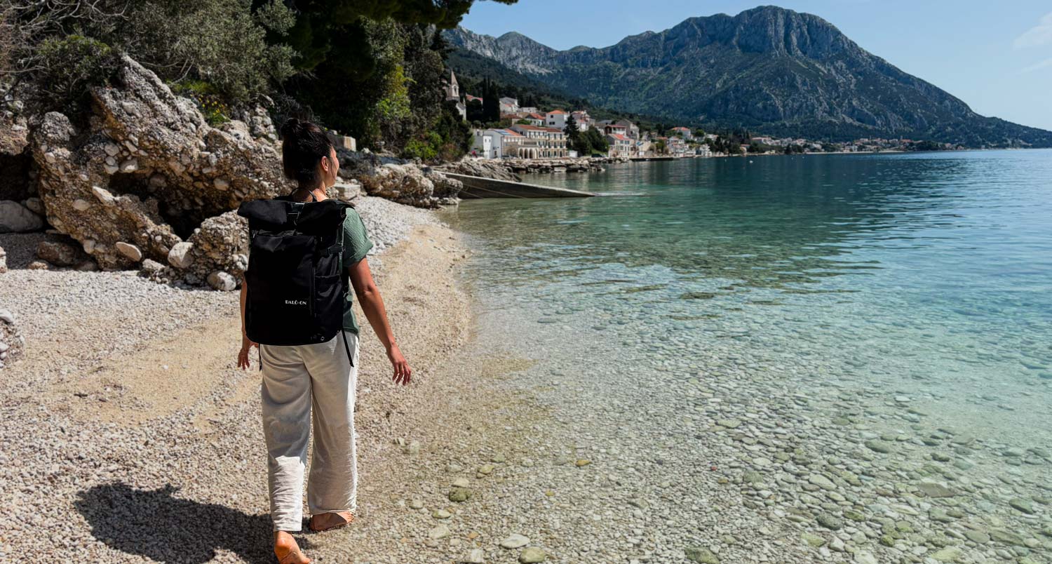 Junge Frau mit schwarzem Rucksack der Marke BALÔ-ON läuft am Strand und blickt in die Ferne; im Hintergrund türkisfarbenes Wasser und Berge.