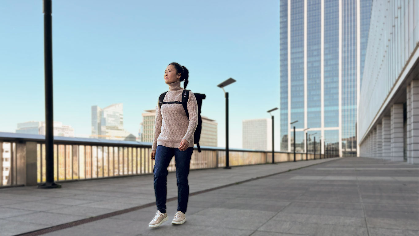 Junge Frau mit schwarzen Rucksack der Marke BALÔ-ON geht über einen Platz in der Stadt; im Hintergrund moderne graue Hochhäuser.
