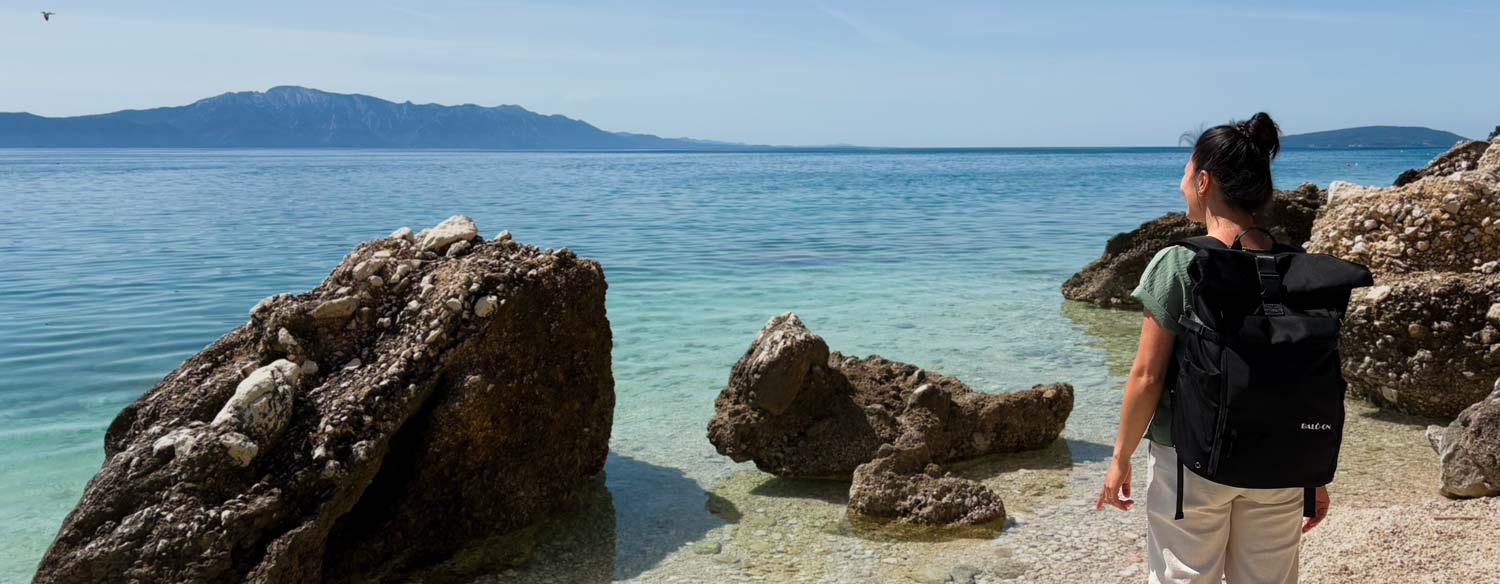 Junge Frau mit schwarzem Rucksack der Marke BALÔ-ON steht am Strand, blickt in die Ferne; türkisfarbenes Wasser, Felsen und Berge im Hintergrund. Bild im Querformat.