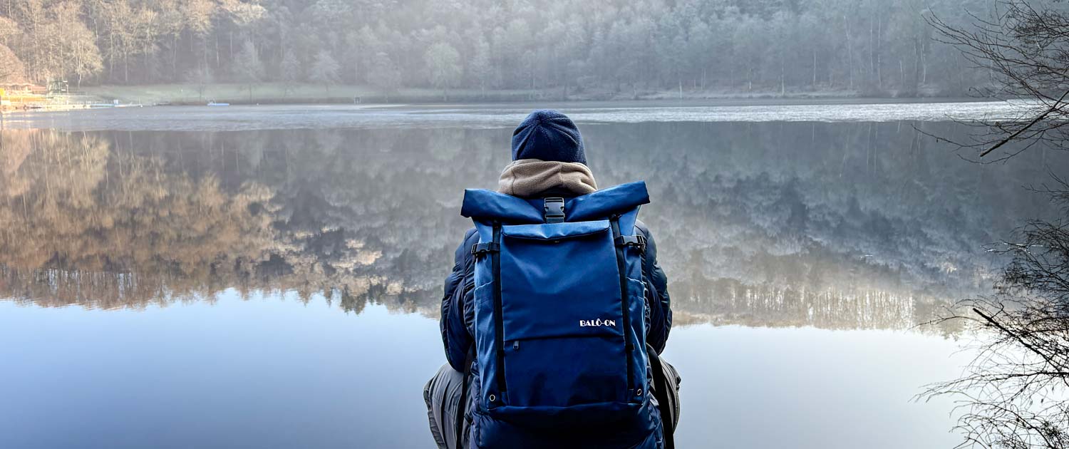 Mann trägt BALÔ-ON Rucksack und hockt am Maar, schaut in die Ferne; im Wasser spiegeln sich Wald und Himmel, herbstlich-winterliche Lichtstimmung.