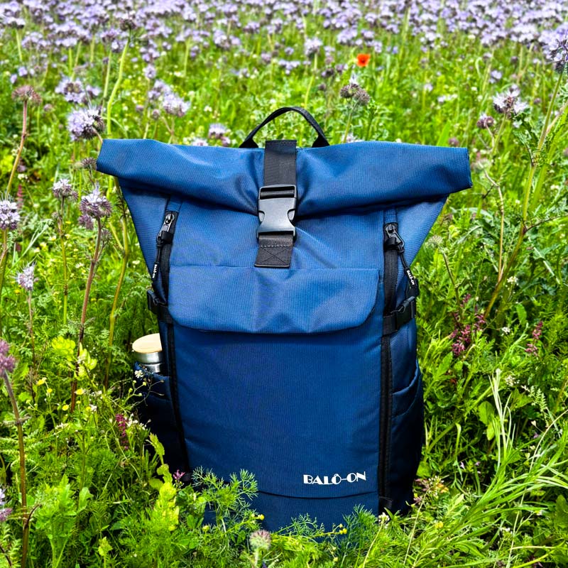 Blauer BALÔ-one-Rolltop-Rucksack steht auf einer bienenfreundlichen Wiese mit lila blühenden Blumen.