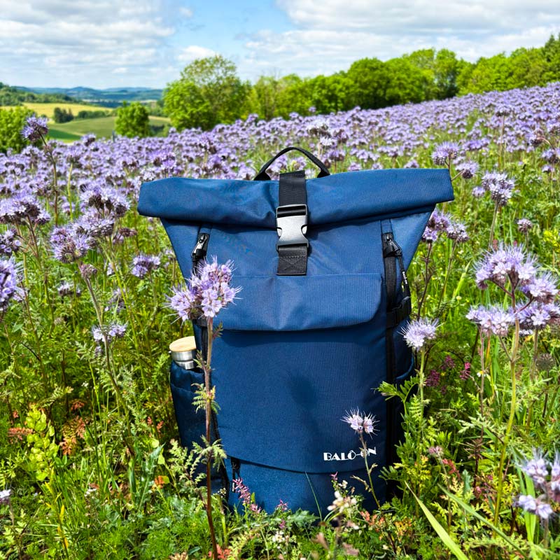 BALÔ-one Blue 25 Liter Rucksack steht auf einer Blumenwiese; im Hintergrund erstreckt sich eine idyllische Landschaft aus Wiesen und Wald