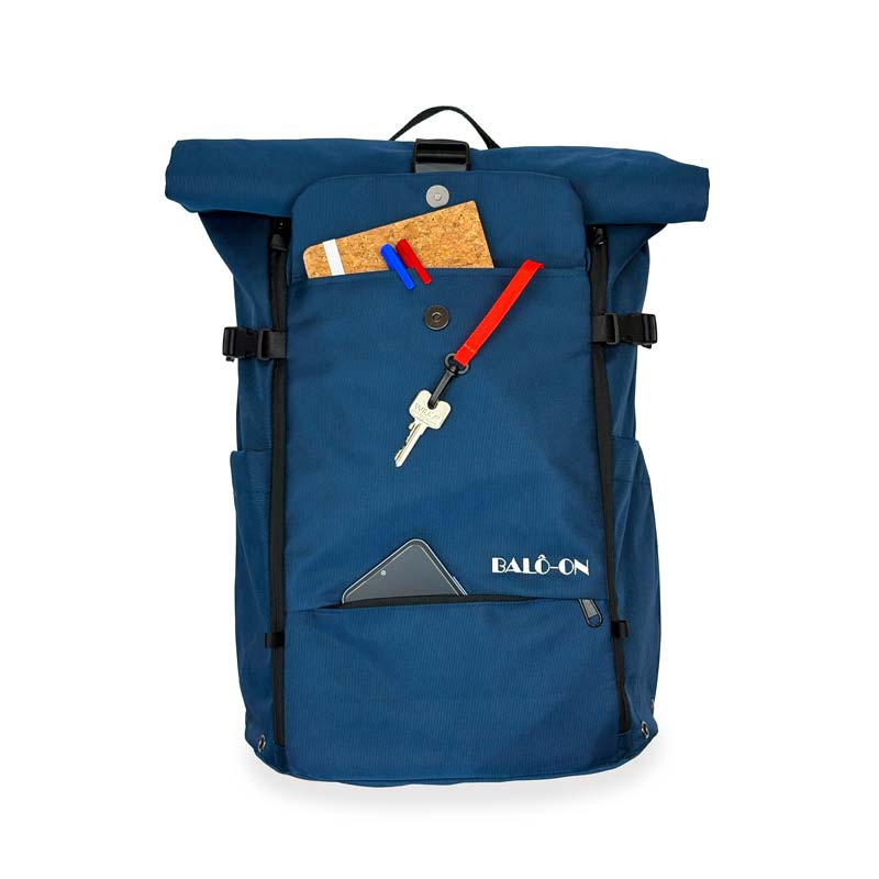 Blauer Rolltoprucksack der marke BALÔ-ON, Vorderansicht; die Fronttasche mit Magnetverschluss ist offen, darin befinden sich Stifte, ein Notizbuch sowie ein Schlüsselanhänger mit rotem Band und Schlüssel. Eine weitere Fronttasche (Reisverschluss) ist geöffnet und mit einem Handy gefüllt.