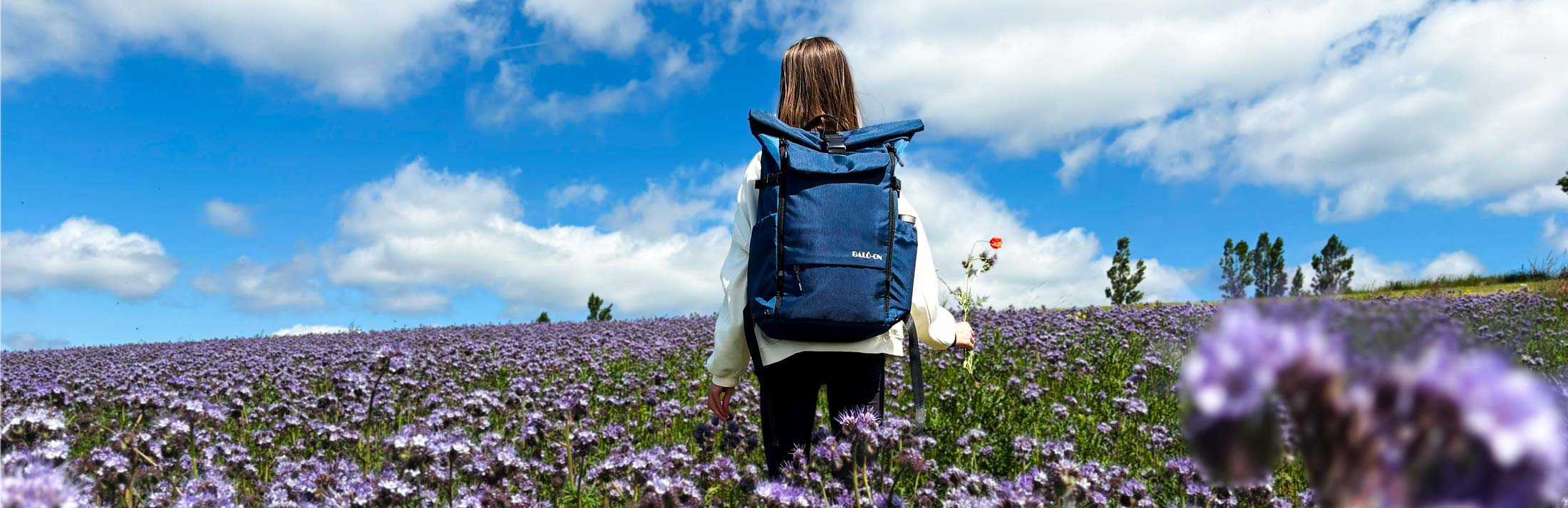 Mädchen mit blauem BALÔ-one Rucksack steht in einer Blumenwiese und hält in der rechten Hand einen kleinen Blumenstrauß.