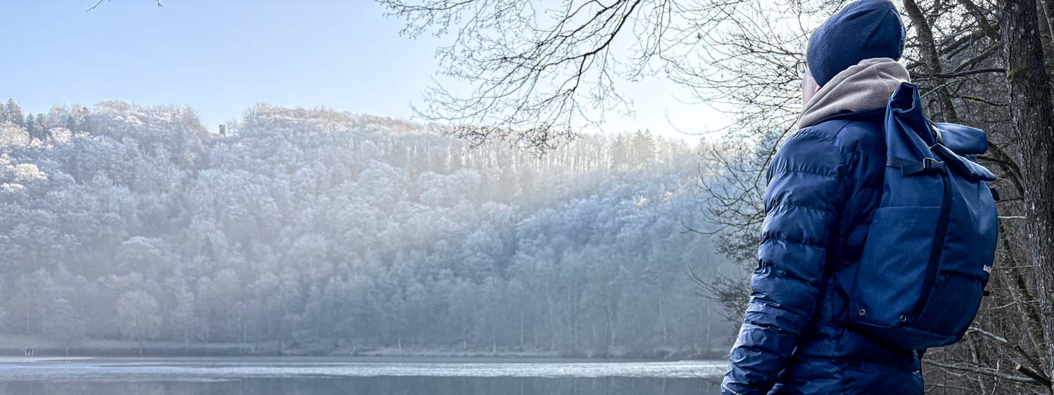Mann trägt BALÔ-ON Rucksack und steht am Maar, schaut in die Ferne; winterliche Lichtstimmung.