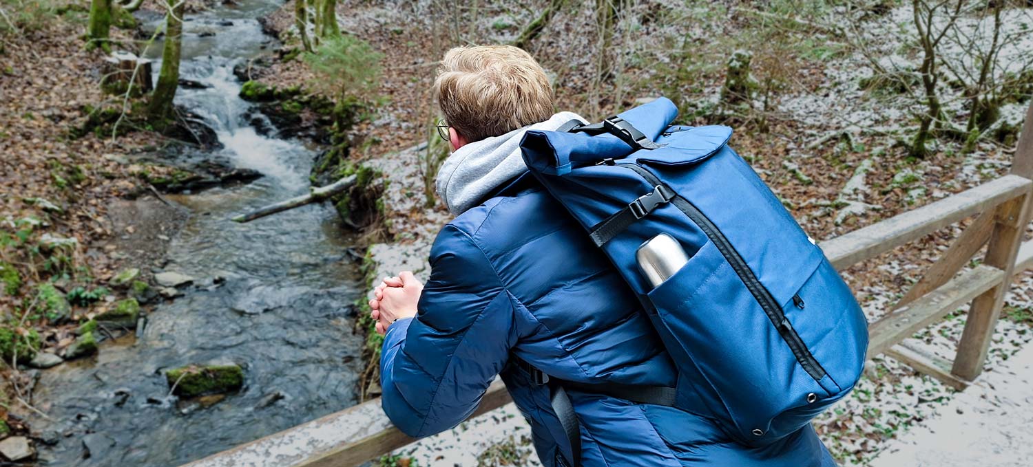 Mann trägt einen BALÔ-one blue 25 L und steht auf einer leicht mit Schnee bedeckten Holzbrücke. Im Hintergrund sind ein Bach und herbstliches Laub zu sehen.
