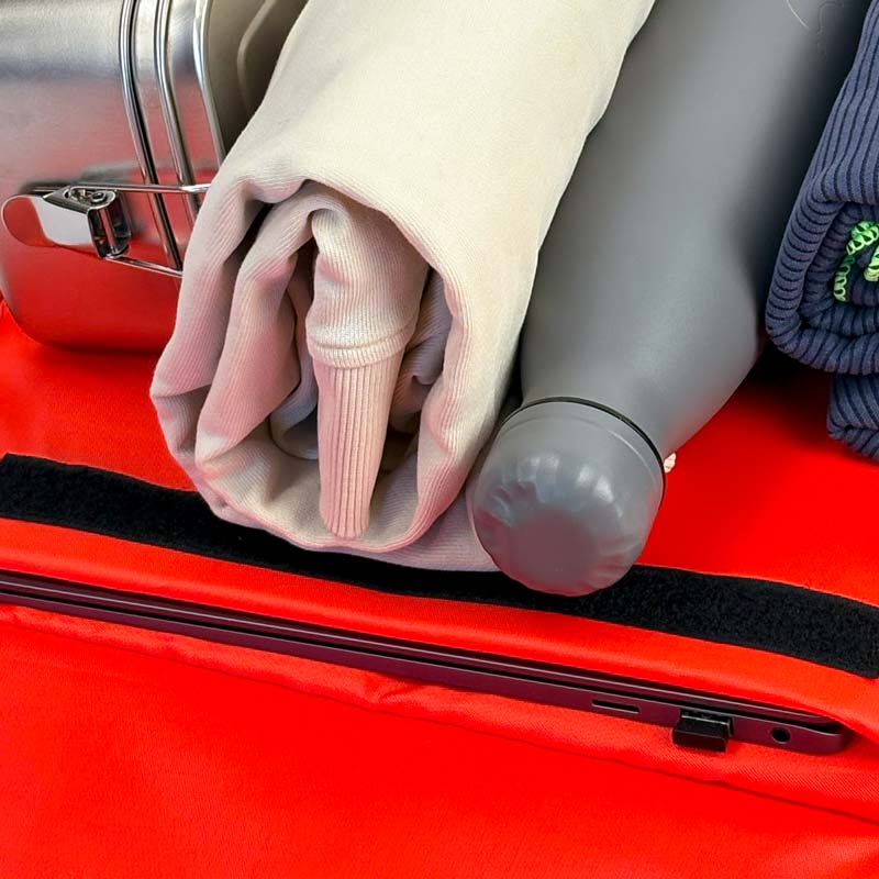 Innenansicht des Hauptfachs und des gepolsterten Laptopfachs; gefüllt mit Laptop, Brotdose aus Edelstahl, Pullover, Trinkflasche und Handtuch. Der Innenstoff ist rot.