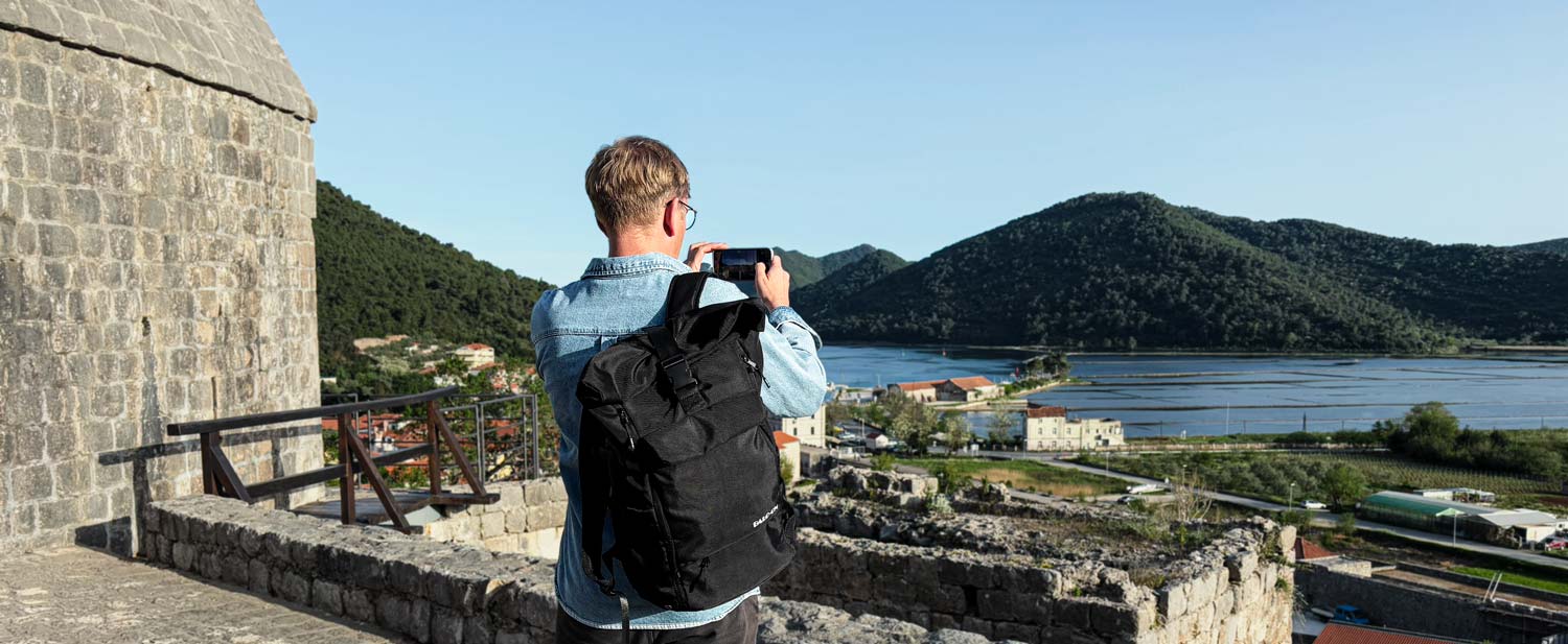 Junger Mann mit BALÔ-ON Rucksack steht auf einer historischen Mauer und fotografiert mit dem Handy die Berglandschaft und den Salzabbau.