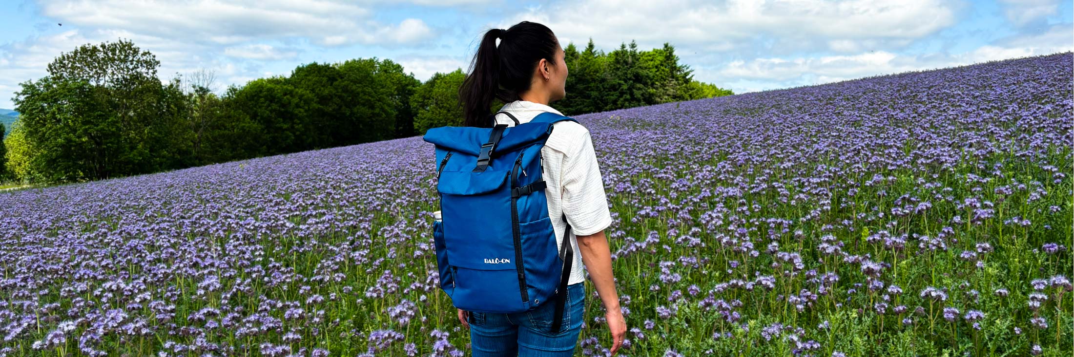 Eine Frau wandert durch ein Blumenfeld und trägt einen blauen Rolltop-Rucksack von BALÔ-ON auf dem Rücken.