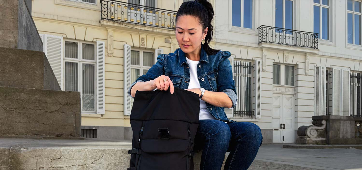 Junge Frau sitzt auf einer Steinmauer und öffnet ihren Rolltop-Rucksack der Marke BALÔ-ON; im Hintergrund sind historische Gebäude zu sehen.