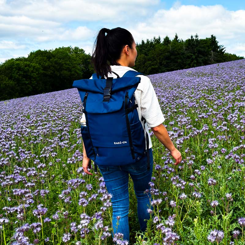 Eine Frau wandert durch ein Blumenfeld und trägt einen blauen Rolltop-Rucksack von BALÔ-ON auf dem Rücken.