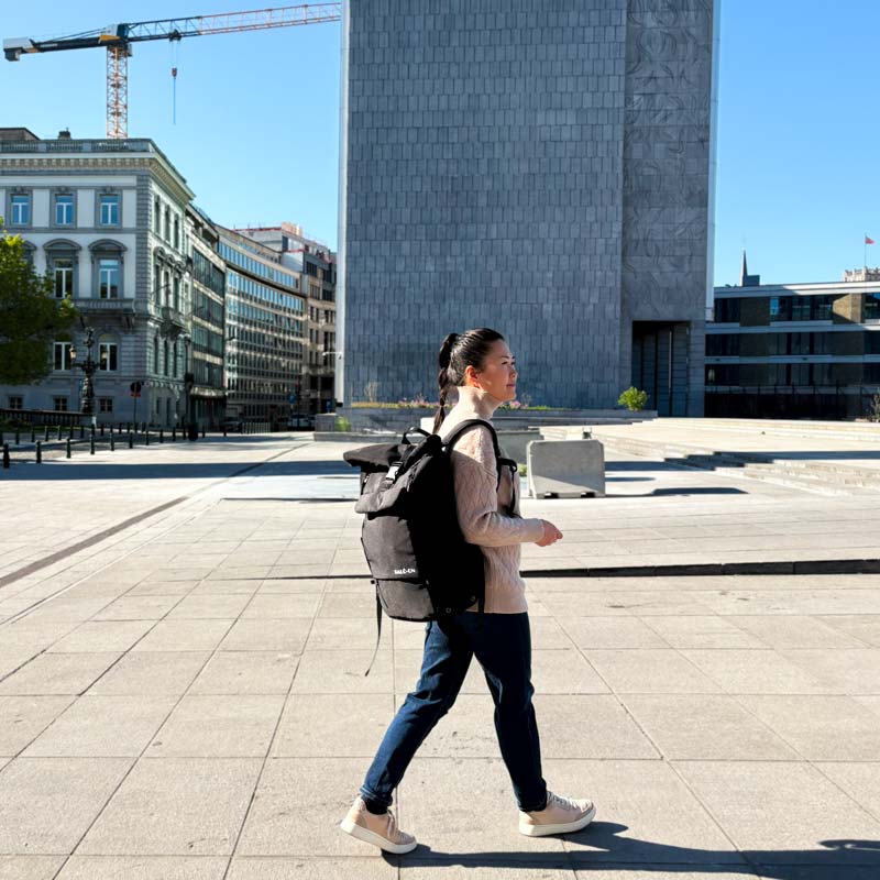 Junge Frau mit schwarzen Rucksack der Marke BALÔ-ON geht über einen Platz in der Stadt; im Hintergrund modernes graues Hochhaus, historische Gebäude, ein Kran und blauer Himmel.
