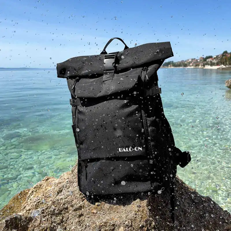 Wasserdichter Rolltop-Rucksack, Farbe schwarz, der Marke „BALÔ-ON“  auf einem Felsen am Meer mit türkisfarbenem Wasser, der von Wasser gespritzt wird und dabei trocken bleibt