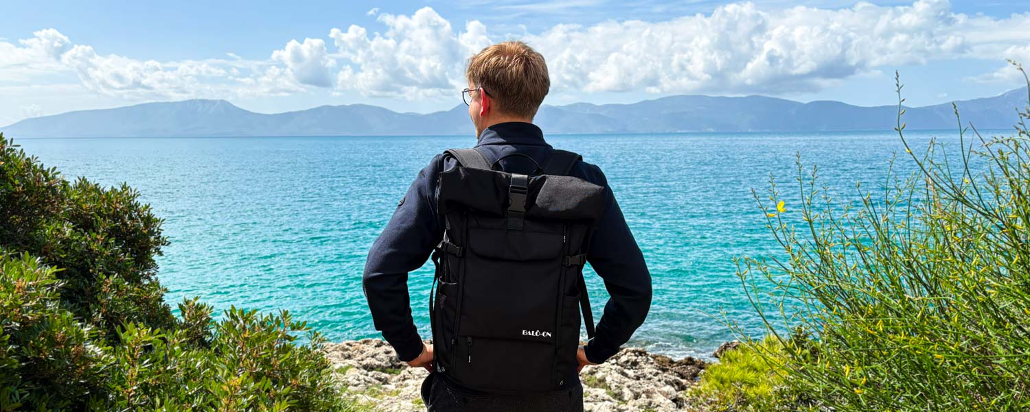 Junger Mann trägt den BALÔ-one Black 25L-Rucksack und schaut auf das offene türkisfarbene Meer; blauer Himmel mit Schäfchenwolken