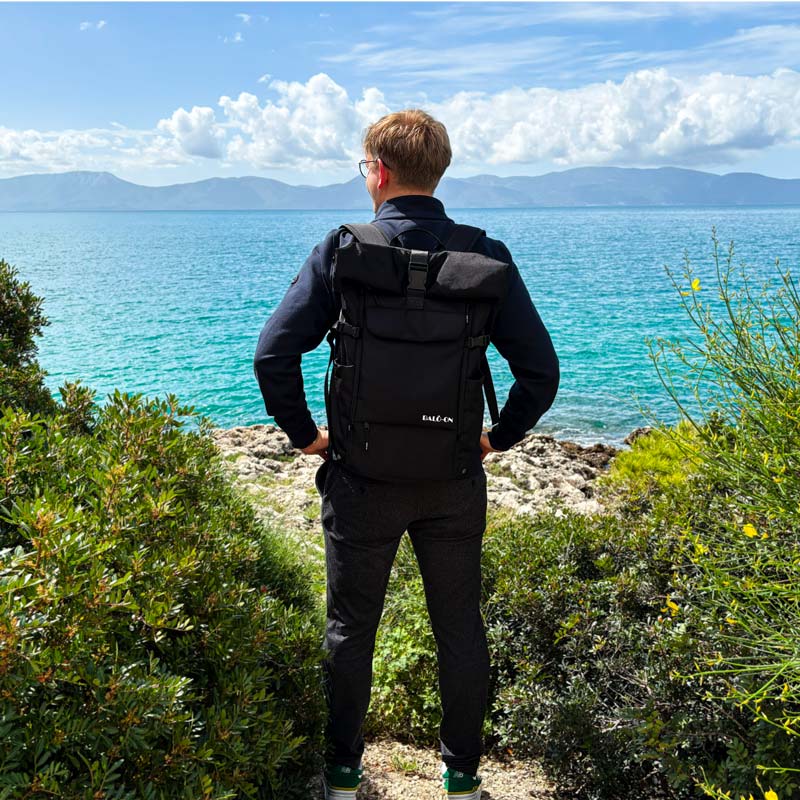 Junger Mann trägt den BALÔ-one Black 25L-Rucksack und schaut auf das offene türkisfarbene Meer; blauer Himmel mit Schäfchenwolken