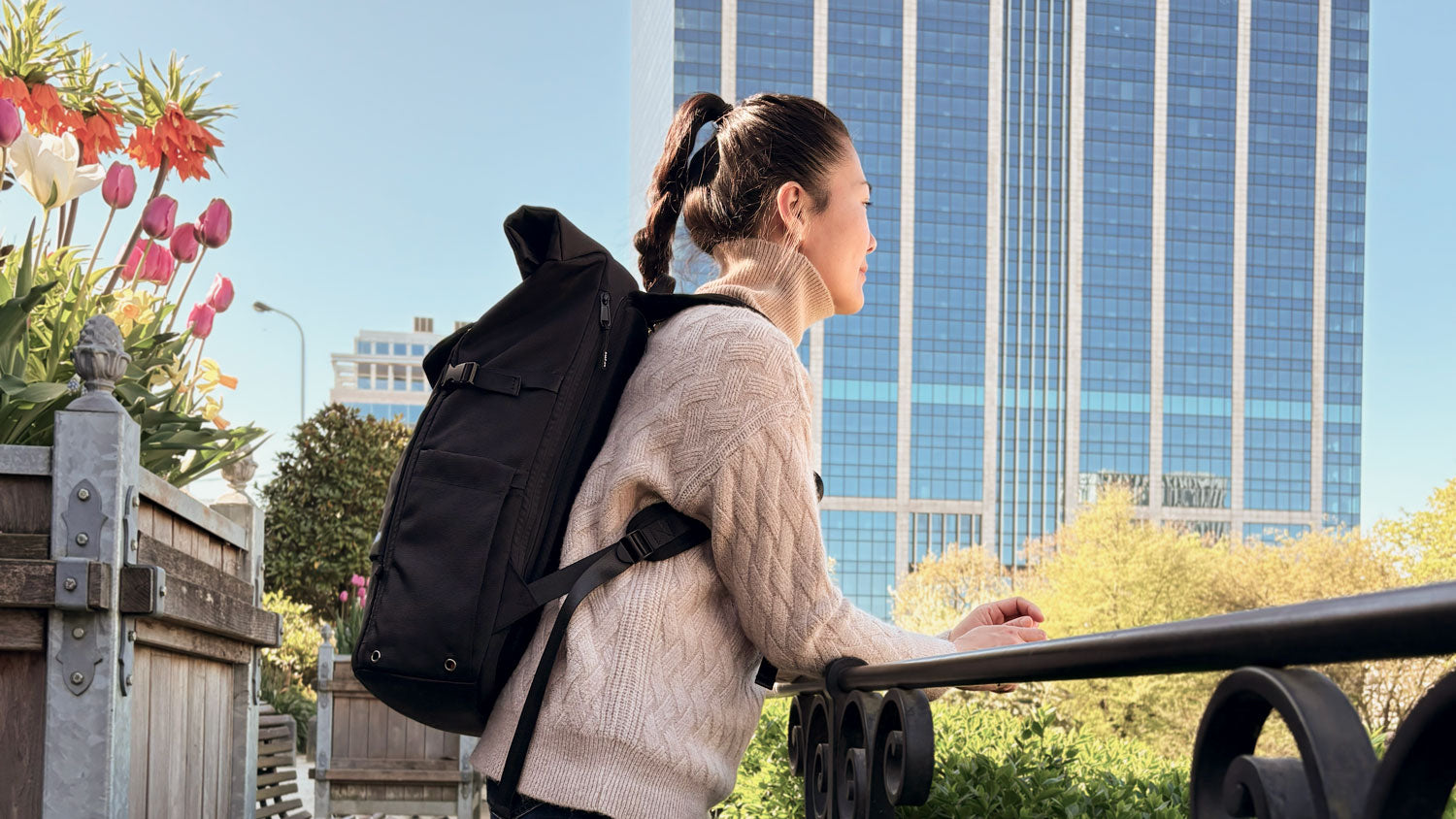 Frau trägt einen schwarzen Rucksack der Marke BALÔ-ON im Park; im Hintergrund ein Hochhaus vor blauem Himmel, im Blumenkübel blühen Frühlingsblumen.

