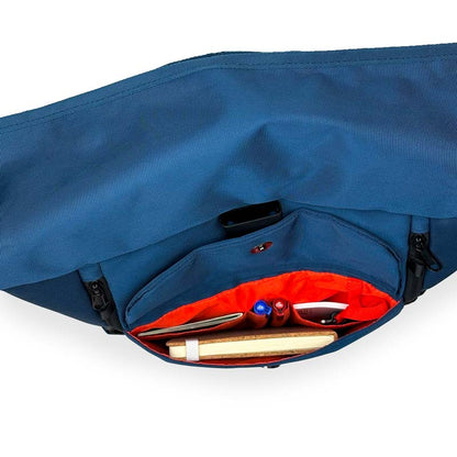 Blauer Rolltoprucksack der Marke BALÔ-ON, Ansicht von oben; geöffnete Fronttasche mit Magnetverschluss und Schlüsselanhänger, gefüllt mit Stiften und Notizbuch.