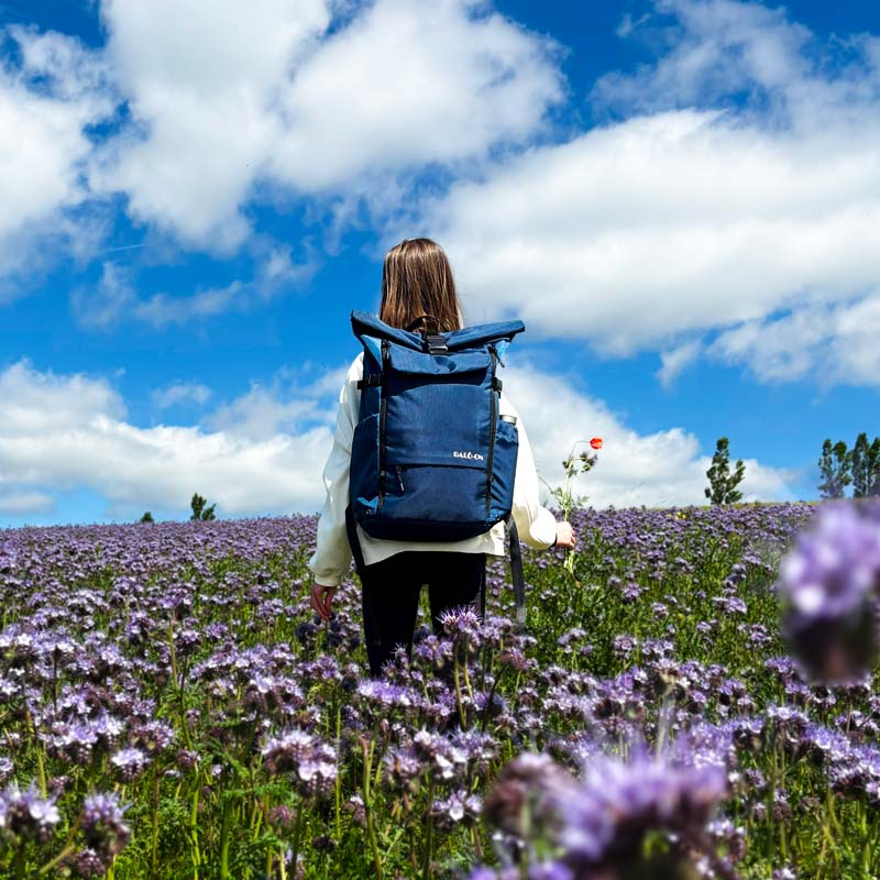 Mädchen mit blauem BALÔ-one Rucksack steht in einer Blumenwiese und hält in der rechten Hand einen kleinen Blumenstrauß.