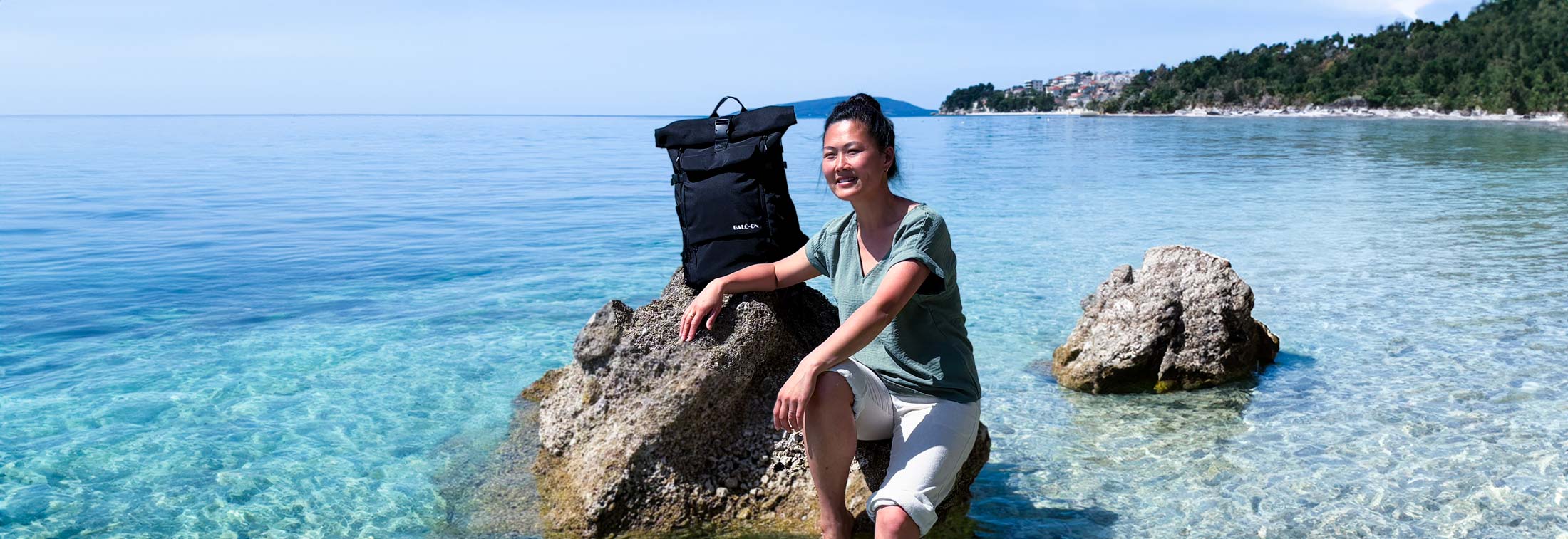 Schwarzer BALÔ-ON Rucksack steht auf einem kleinen Felsen im türkisfarbenen Meer; daneben sitzt eine junge Frau mit den Füßen im Wasser. Im Hintergrund sind eine kleine Ortschaft und ein Kiefernwald entlang des Strandes zu sehen.